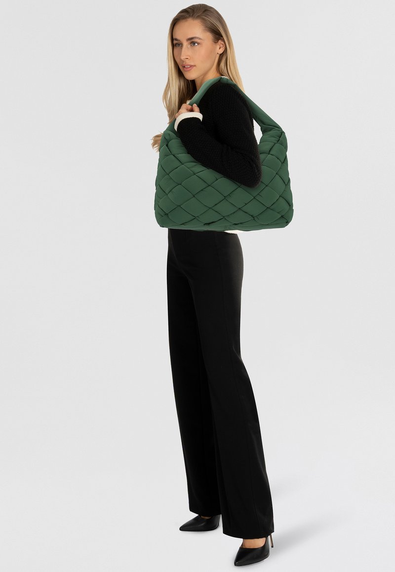 Sac à bandoulière vert matelassé avec une large sangle douce, présentant une surface texturée et une forme décontractée. Porté avec un pantalon et des chaussures noires.