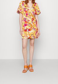 Robe fleuri en jaune, rose et orange avec un design en portefeuille et des manches courtes bouffantes. Associée à des sandales à talons carrés orange.