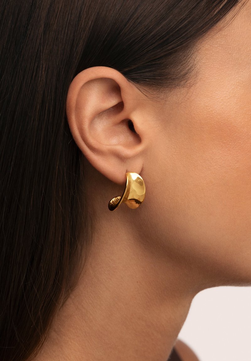 Pendientes de oro con un diseño suave y curvado. Presentan un acento texturizado y un acabado pulido, exhibidos en la oreja de un modelo.