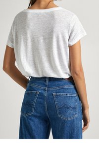 Osoba oblečená v volné bílé krátké tričko zasunuté do vysokých modrých jeansů z denimu s zadními kapsami a jemnou výšivkou.