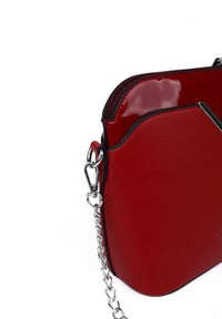 Červená kabelka z umělé kůže s lesklým povrchem, na zip a se stříbrným řetízkem jako uchem. Zakřivený tvar s ostrými akcenty poblíž zipu.