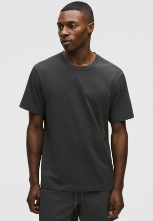 Junger Mann mit dunkler Haut, der ein schlichtes dunkelgraues kurzärmeliges T-Shirt und passende Hosen trägt, vor einem hellgrauen Hintergrund stehend.