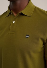 Olivgrön polo skjorta i texturerad tyg med en tre-knappsknäppning och en svart-vit broderad logotyp på bröstet.