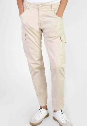 Pantalon cargo beige avec poches latérales porté par une personne les mains dans les poches, associé à des baskets blanches sur un fond uni.