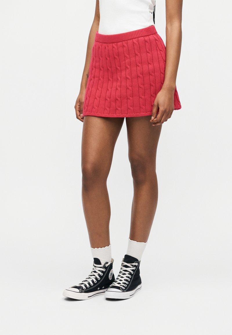 Γυναίκα που φοράει κοντή κόκκινη φούστα πλεκτή με σχέδιο καλώδιο, λευκές κάλτσες, μαύρα ψηλά αθλητικά παπούτσια και λευκό αμάνικο τοπ, όρθια σε λευκό φόντο.