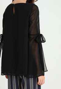 Svart, genomskinlig blus med långa puffärmar, prydd med knytaccenter vid ärmsluten och en nyckelhålsöppning på baksidan. Stylande med randiga byxor.