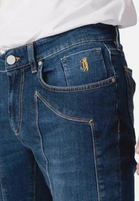 Jeans in denim blu scuro con cuciture dorate, dotati di una tasca frontale e un piccolo design ricamato vicino alla tasca.