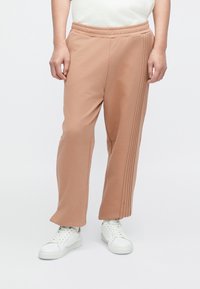 Vaaleanruskeat collegehousut, joissa on resorivyötärö, sivuilta laskostettu yksityiskohta ja resorit lahkeensuissa, yhdistetty valkoisiin tennareihin. Sileä ja pehmeä pinta.