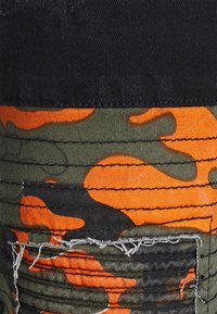 Tela con patrón de camuflaje que presenta tonos naranjas, negros y verdes, con textura cosida. Bordes deshilachados y un detalle de bolsillo visible.