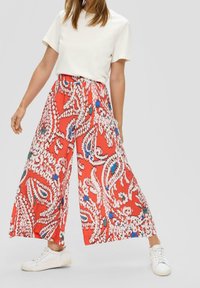 Rote weit geschnittene Hosen mit einem Paisleymuster in Weiß und Blau. Kombiniert mit einem schlichten cremefarbenen T-Shirt und weißen Turnschuhen.