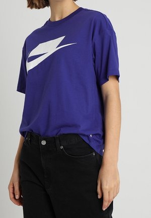 Osoba nosząca luźny fioletowy T-shirt z dużym białym stylizowanym logo, lekko wpuszczonym w czarne jeansy z wysokim stanem.