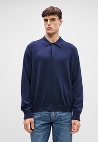 Ναυτρίκης μπλε πλεκτή μπλούζα με γιακά πόλο, μακριά μανίκια και κουμπιά. Ριμπέ μανσέτες και στρίφωμα. Φοράει με ανοιχτό μπλε τζιν.