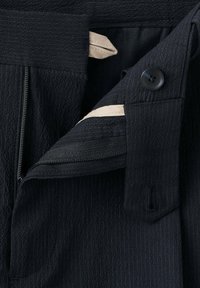 Gros plan d’un pantalon sombre texturé non fermé avec bouton et doublure intérieure beige visible près de la ceinture.