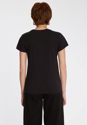 Frau mit kurzem braunem Haar, die ein schlichtes schwarzes T-Shirt mit kurzen Ärmeln und schwarze Hose trägt, von hinten vor einem schlichten weißen Hintergrund gezeigt.