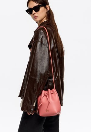 Różowa skórzana torba typu bucket z zamknięciem na sznurek i złotym akcentem z logo, noszona na brązowej skórzanej kurtce na białym tle.