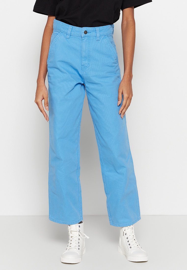 Pantaloni larghi azzurri chiari realizzati in tessuto di tela, dotati di chiusura con bottone, passanti per cintura e una texture liscia. Abbinati a sneaker bianche alte.