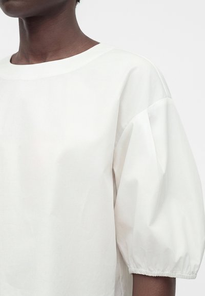 Άτομο με σκουρόχρωμο δέρμα που φοράει λευκή χαλαρή μπλούζα με στρογγυλή λαιμόκοψη και φουσκωτά μανίκια που τελειώνουν σε ελαστικά μανσέτα.