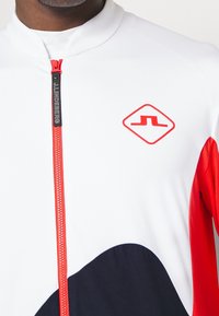 Vit idrottstopp med röda accenter, dragkedja vid kragen och svart böjd design. Har en logo i röd diamantform och textil med struktur.