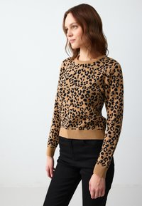 Sweter w cętki z leopardem w kolorze beżowym i czarnym, wykonany z dzianiny, z okrągłym dekoltem oraz ściągaczami przy rękawach i u dołu. W połączeniu z czarnymi spodniami.