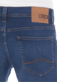 Blue denim jeans featuring a brown leather patch labeled "MUSTANG True Denim" and visible orange stitching along the pockets and seams.