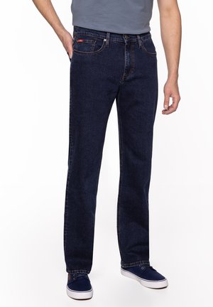 Mężczyzna w granatowych prostych jeansach z przednimi kieszeniami i granatowymi trampkami, stojący na białym tle.
