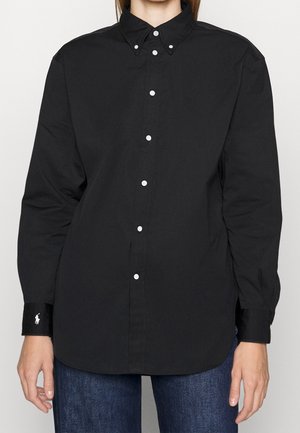 Personne portant une chemise noire à manches longues boutonnée avec des boutons blancs et un jean bleu foncé, debout devant un fond uni.