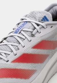 Grijze sportschoen met rode accenten, gestructureerde bovenkant, witte veters en blauw logo; heeft een slank ontwerp en een gevoerde zool.