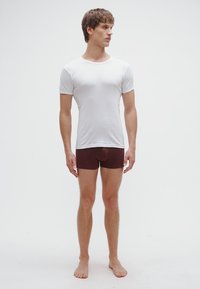 Witte nauwsluitende katoenen t-shirt gecombineerd met bordeauxrode stretchshorts, getoond op een staande mannelijke model, blootsvoets op een effen achtergrond.