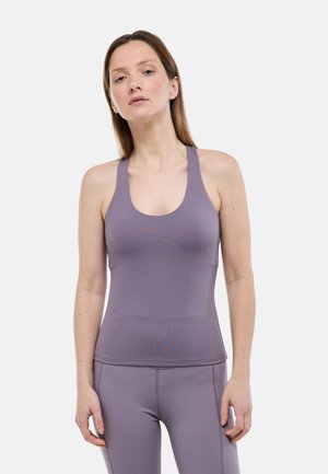 Femme portant un débardeur de sport violet et un legging, debout avec les bras détendus devant un fond clair uni.