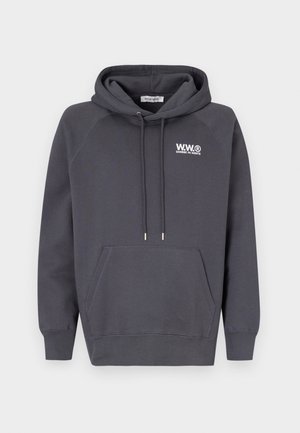 Henkilö, jolla on yllään tummanharmaa huppari, jossa on logo, tummat farkut, valkoiset lenkkarit ja musta olkalaukku, yksiväristä vaaleaa taustaa vasten.