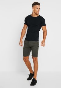 Camiseta negra ajustada de manga corta, combinada con pantalones cortos verde olivo con bolsillos laterales, llevada con zapatillas negras. Diseño simple y casual.