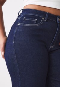 Gros plan d’une personne portant un jean en denim bleu foncé taille haute avec des coutures visibles et une petite poche, montrant une partie de son ventre.