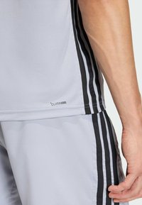 Bočný pohľad na osobu v svetlosivých športových šortkách a tričku s čiernymi tromi pruhmi a štítkom „Climacool“ na leme trička.