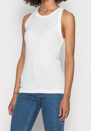 Frau trägt ein ärmelloses weißes Tanktop und blaue Jeans, steht vor einem schlichten hellen Hintergrund.