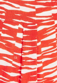 Röd och vit tyg med abstrakt tigerstripesmönster och liten "ck"-logotyp broderad nära en vertikal söm.