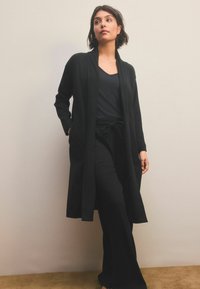 Sort ribbet lang cardigan med åben front og sidelommer, parret med en tætsiddende sort top og vide bukser, alt sammen i et blødt strikket materiale.