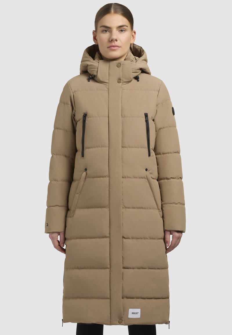 Abrigo largo beige tipo plumón con capucha, diseño acolchado, bolsillos con cremallera negra y un parche de logo en el dobladillo. Textura suave y corte holgado.