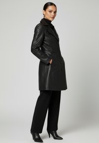 Kvinna står i profil iklädd en svart läderrock med knappar, svarta byxor och svarta stövlar med klackar mot en enkel ljusgrå bakgrund.