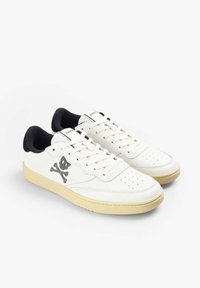 Zapatillas de cuero blancas con acentos en negro, con perforaciones para transpirabilidad y un diseño de calavera y huesos cruzados en plata. Suela de goma.