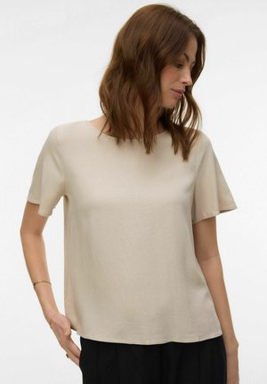 Žena sa smeđom kosom srednje dužine, odjevena u bež majicu kratkih rukava i crne hlače, gleda prema dolje na svoju desnu stranu.