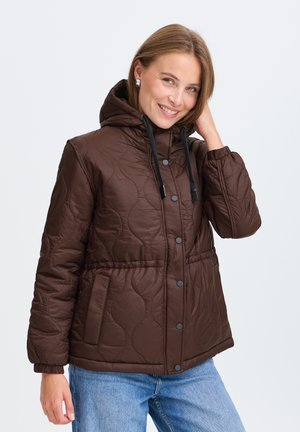 Femme souriante portant une veste matelassée marron à capuche avec des cordons noirs et un jean bleu, posant avec une main touchant sa tête.