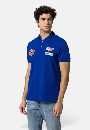 Junger Mann mit lockigem Haar, der ein blaues Polohemd mit den Aufnähern "1969" und "Grand Prix" trägt, kombiniert mit hellblauen Jeans, blickt nach rechts.