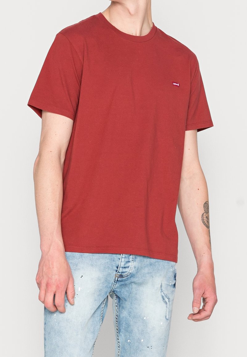 Homme portant un t-shirt rouge uni à manches courtes avec un petit logo sur la poitrine, associé à un jean bleu clair déchiré.