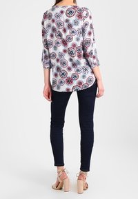 Langærmet bluse med et blomster mandala mønster i rød, blå og hvid, parret med mørke denim jeans og stripsandaler i beige.