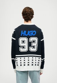 Mornarski pulover s krojem okroglega izreza, narejen iz pletene tkanine, z belimi dodatki, zlogom "HUGO" v modri barvi in velikim "93" na hrbtu s vzorčastim robom.