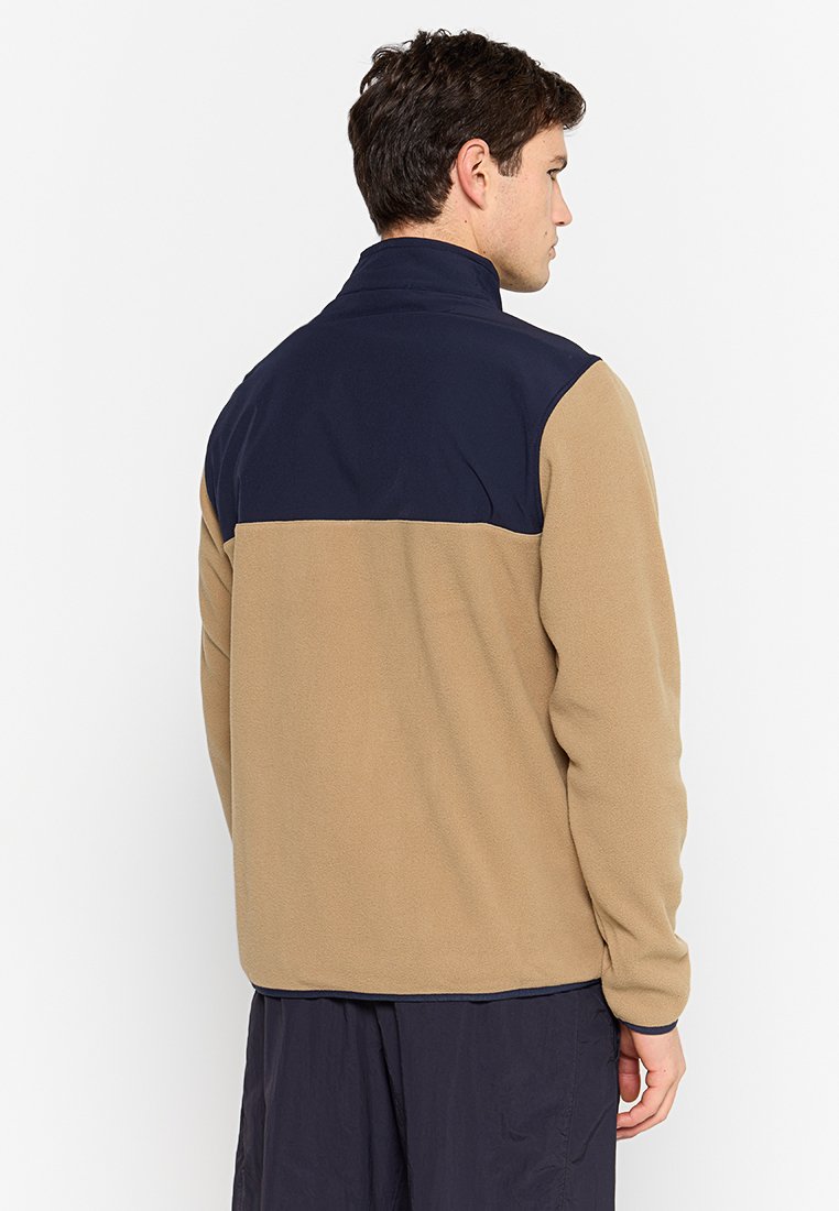 Homme portant une veste en polaire beige et marine, tourné le dos contre un fond blanc uni.