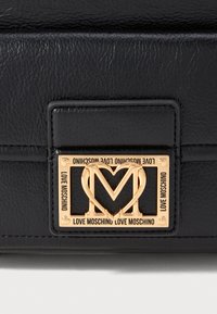 Juodas odinis krepšys su auksiniu stačiakampiu rėmu, kuriame yra širdies formos "M" ir visose pusėse esantis "Love Moschino" tekstas.