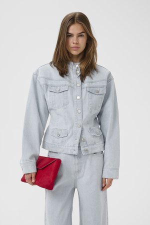 Žena v svetlomodrej rifľovej bunde a džínsoch drží červenú listovú kabelku na jednoliatom pozadí.