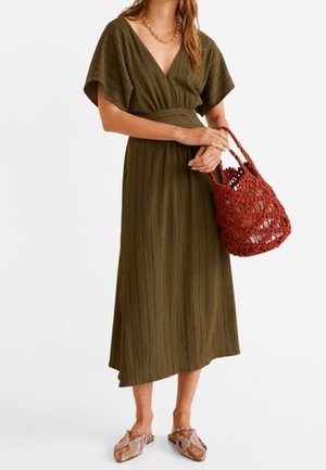 Kobieta w oliwkowej, midi sukience z dekoltem w kształcie litery V i szerokimi rękawami, trzymająca czerwoną plecioną torebkę i nosząca wzorzyste buty wsuwane.