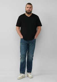 Schwarzes V-Ausschnitt-T-Shirt, blaue Jeans und weiße Sneakers. Das Model steht mit den Händen in den Taschen vor einem neutralen grauen Hintergrund.